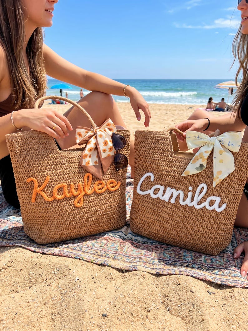 May include: Two woven straw tote bags featuring embroidered names. The left bag displays the name Kaylee in orange thread with a floral scarf, while the right bag shows the name Camila in white thread with a lemon-patterned scarf attached to the handle.