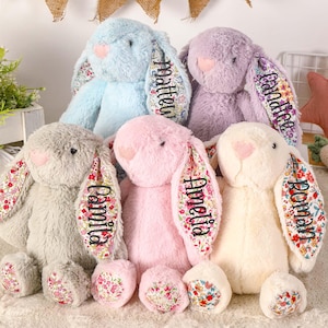 以下が含まれることがあります： 花柄の生地の耳が付いた、ぬいぐるみ製のウサギの玩具が5つあります。ウサギは、薄い青色、紫色、ピンク色、灰色、クリーム色と、さまざまな色をしています。それぞれのウサギの耳には、名前が刺繍されています。名前は、マシュー、キャンディス、アメリア、カミラ、ローマンです。