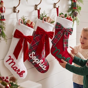 May include: Three Christmas stockings hanging on hooks. The first is white with a red velvet bow and the name Mark. The second is red and green plaid with a red velvet bow and the name Sophia. The third is red with a plaid bow and the name Noah.