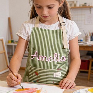 以下が含まれることがあります： ピンク色の筆記体で「Amelia」と書かれた、緑と白のストライプのエプロン。エプロンにはポケットがあり、水彩絵の具で絵を描いている人が着用しています。エプロンは丈夫な生地で作られています。