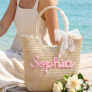 May include: A beige woven straw tote bag featuring the name Sophia in raised pink script lettering on the front. The bag has two sturdy handles, one of which is adorned with a white lace ribbon tied into a bow, providing a textured appearance.