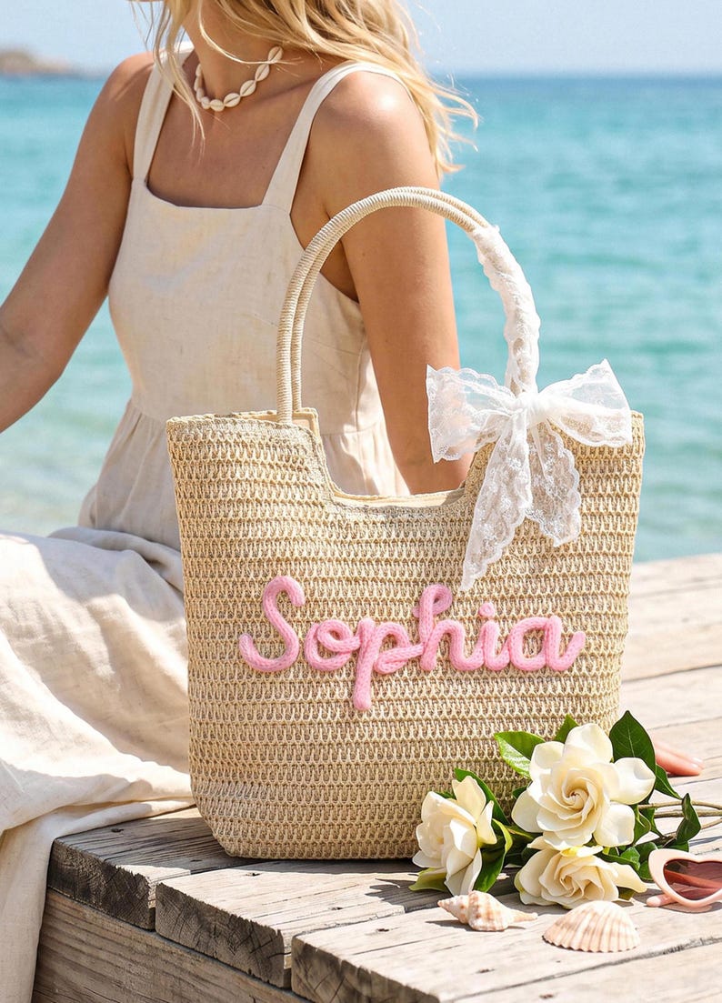 May include: A beige woven straw tote bag featuring the name Sophia in raised pink script lettering on the front. The bag has two sturdy handles, one of which is adorned with a white lace ribbon tied into a bow, providing a textured appearance.