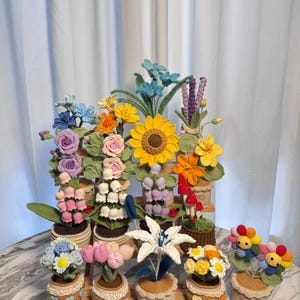 Könnte beinhalten: Eine Sammlung bunter, gehäkelter Blumen in kleinen Töpfen. Die Blumen, darunter Sonnenblumen und Rosen, sind auf einem Holzregal angeordnet. Die Töpfe haben eine dekorative Spitzenborte.