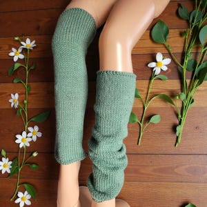 Op de afbeelding: Een paar saliegroene gebreide beenwarmers. Eén warmer is tot aan de knie opgetrokken, de andere is opgerold. De beenwarmers worden getoond op een mannequinbeen. Witte bloemen en groene bladeren zijn op een houten oppervlak geplaatst.
