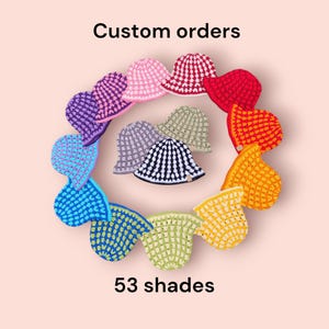 May include: A selection of crocheted hats in a rainbow of colours, displayed in a circular arrangement. The hats feature shades of pink, red, orange, yellow, green, blue, purple, and black and white. The text "Custom orders" and "53 shades" are visible.