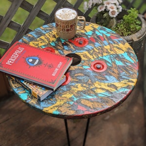 May include: A colorful, round wooden table with a single black metal leg. The table top has a unique, abstract design with red, yellow, blue, and green paint. Two books are stacked on the table, one red and one blue. A brown and white coffee mug is on the table.