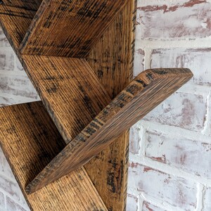 May include: Rustic wooden shelf unit with a staggered design. The shelf is made of reclaimed wood with a dark brown stain and a distressed finish. The shelf is mounted on a white brick wall.