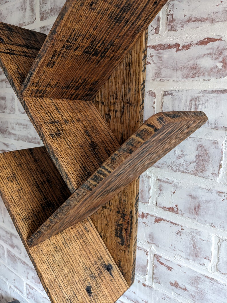 May include: Rustic wooden shelf unit with a staggered design. The shelf is made of reclaimed wood with a dark brown stain and a distressed finish. The shelf is mounted on a white brick wall.