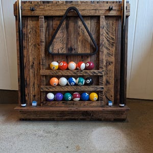 May include: A rustic wooden pool ball rack with a triangle rack and a set of billiard balls. The rack is made of reclaimed wood and has two shelves to hold the balls. Two pool cues are on either side.