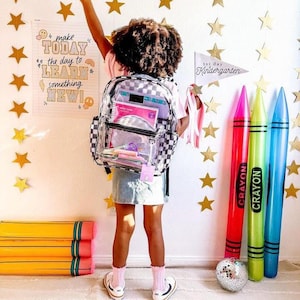 May include: A clear backpack with a checkered pattern and a variety of school supplies inside. The backpack is being worn by a young person with long, dark hair. The background is a white wall with gold stars and a sign that says "Make Today the Day to Learn Something New!"