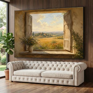 May include: A large rectangular wall art print depicting an open window with wooden shutters overlooking a golden countryside landscape. A ceramic vase with wildflowers sits on the stone windowsill. Below, a cream-colored tufted sofa is positioned.