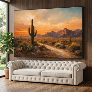 May include: A large rectangular wall art print depicting a desert landscape at sunset with a tall saguaro cactus in the foreground, a winding dirt path, and distant mountains under a vibrant orange and yellow sky, mounted above a cream tufted sofa.