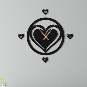 Puede incluir: Reloj de pared negro en forma de corazón con marcadores de hora en números romanos y manecillas doradas. El reloj está montado en una pared blanca sobre una cómoda blanca con dos cajones. Una pequeña planta en maceta está sobre la cómoda.