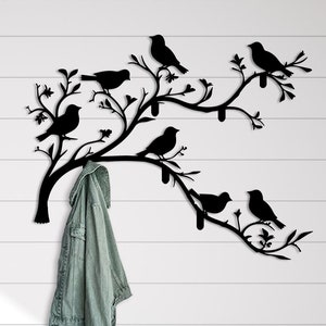 May include: Black metal wall-mounted coat rack shaped like a tree branch with birds. The rack has a single hook and is mounted on a white wall. A blue jacket is hanging on the hook.