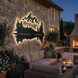 May include: A black metal wall art piece depicting a mountain range, trees, and a lake scene. The art is illuminated with warm white lights, mounted on a stone wall. The outdoor setting includes patio furniture and potted plants.