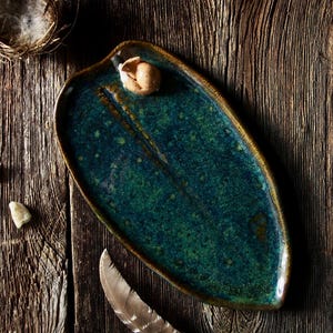 May include: A handmade, teal-colored ceramic serving platter with a leaf-like shape. The platter has a speckled, glossy finish and a raised edge. A small, tan-colored object sits on the platter. The background is a rustic wooden surface.