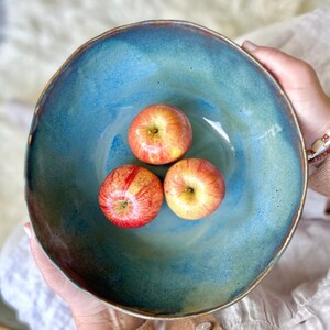 May include: A handmade, ceramic bowl with a blue and green glaze, holding three red and yellow apples. The bowl has a rustic, organic shape and a textured rim. The apples are round and have a vibrant color.