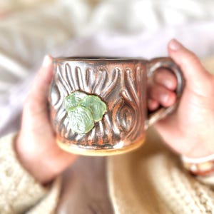 May include: A handmade ceramic mug with a brown and orange textured exterior. The mug features a green, leaf-shaped embellishment. The mug is held by two hands. This unique mug is perfect for enjoying coffee, tea, or other beverages.