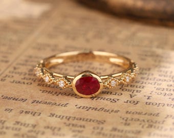 May include: A gold ring with a red ruby gemstone surrounded by small diamonds. The ring is set on a textured, brown surface.