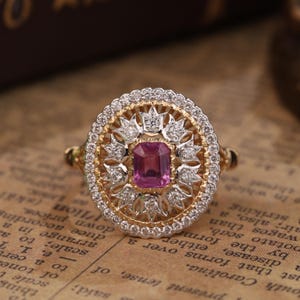 Puede incluir: Un anillo de oro y plata con una gran piedra preciosa rosa rodeada de diamantes más pequeños. El anillo tiene un diseño delicado e intrincado.