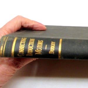 Puede incluir: Un libro antiguo titulado "Connecting Induction Motors" de Dudley. El libro tiene una cubierta verde oscuro con letras y detalles dorados. El libro está sostenido por una mano sobre un fondo blanco.