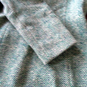May include: A coat featuring a teal and white herringbone pattern. The fabric has a textured, woven appearance with a distinct V-shaped weave throughout the garment. The sleeve is folded over, showing the consistent pattern across the entire surface.