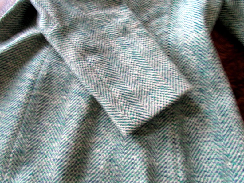 May include: A coat featuring a teal and white herringbone pattern. The fabric has a textured, woven appearance with a distinct V-shaped weave throughout the garment. The sleeve is folded over, showing the consistent pattern across the entire surface.
