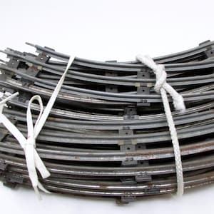 May include: A stack of curved metal train tracks, bound together with a white rope and a white ribbon. The tracks are a dull silver color with some rust visible. The tracks are designed to create a circular or curved layout.