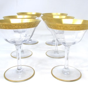 May include: A set of six clear champagne coupe glasses, each with a gold rim and decorative gold band. The glasses feature a classic design with a wide bowl and a slender stem, ideal for serving champagne or cocktails.