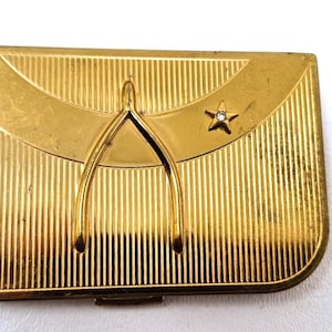 May include: A vintage gold-toned compact with a vertical ribbed design. It features a wishbone and a star with a small gem. The compact has a rectangular shape with rounded edges, suggesting a classic design.