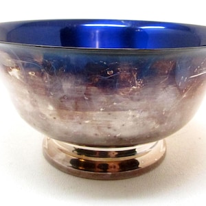 May include: A silver-toned bowl with a vibrant blue interior. The bowl has a wide, slightly flared rim and sits on a round, pedestal base. The interior color contrasts with the exterior, creating a striking visual effect. The bowl appears to be made of metal.