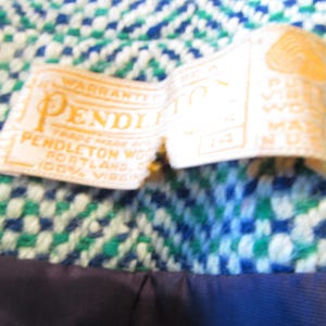 May include: A Pendleton brand clothing label stitched onto a blue, green, and white woven fabric. The white tag features gold text reading Pendleton, Portland, Oregon, 100% virgin wool, and the size 14. A dark blue lining is visible at the bottom.