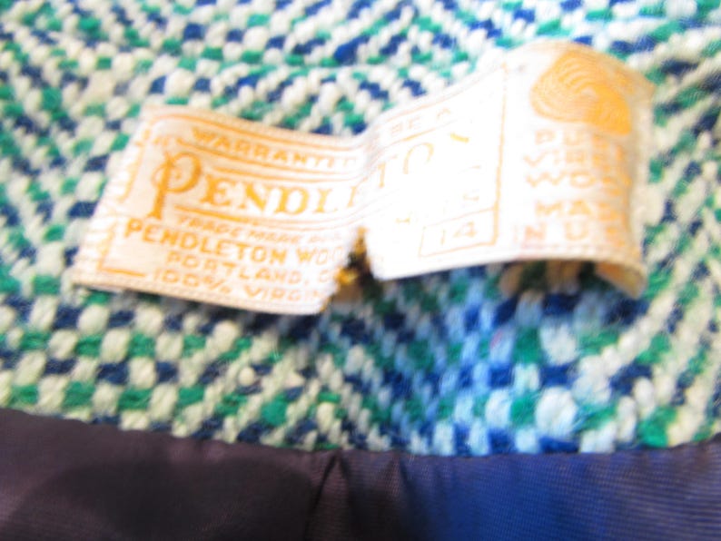 May include: A Pendleton brand clothing label stitched onto a blue, green, and white woven fabric. The white tag features gold text reading Pendleton, Portland, Oregon, 100% virgin wool, and the size 14. A dark blue lining is visible at the bottom.