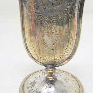 May include: A silver-plated metal vessel with a wide, curved rim and a pedestal base. The body features an embossed floral garland and vertical line pattern with visible tarnish and gold-toned highlights throughout the metallic surface.