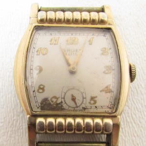 May include: A vintage gold-toned wristwatch with a rectangular face. The watch face is off-white with gold numerals and hands. The brand name "Gruen" is visible. The watch has a small seconds dial and a textured band.