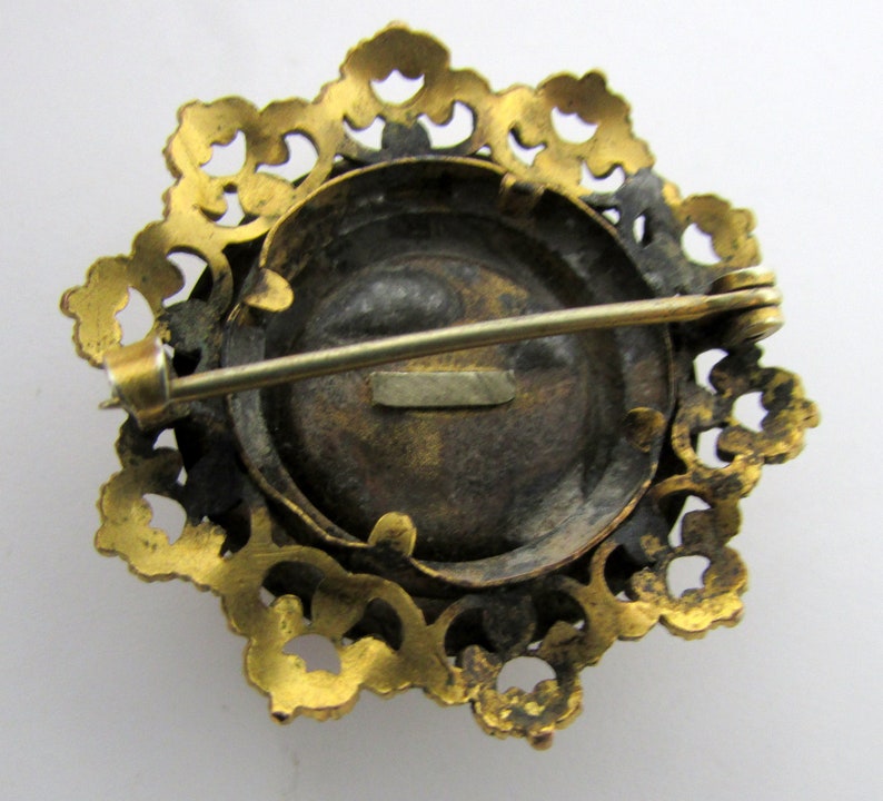 May include: Gold-toned brooch with a round, black cameo set in a filigree frame. The brooch has a pin and catch closure on the back.
