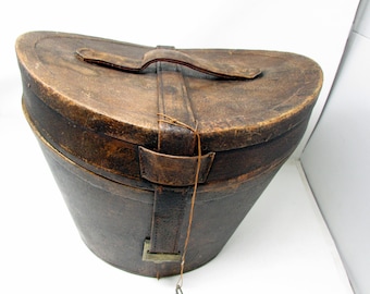 May include: A dark brown, oval-shaped leather hat box with a curved lid and a top handle. A vertical leather strap secures the lid, featuring a metal buckle at the base and a thin, dangling string with a small metal key attached to the front.