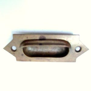 May include: A vintage brass drawer pull with a rectangular shape and a recessed handle. The pull has a decorative design with pointed ends and two circular holes for mounting. The brass has a slightly aged patina.