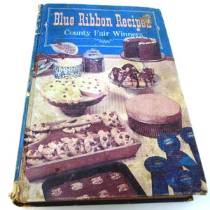 Op de afbeelding: Een vintage kookboek getiteld "Blue Ribbon Recipes County Fair Winners" met een blauwe omslag. De omslag toont afbeeldingen van gebakken goederen, waaronder cakes, taarten en koekjes. Het boek heeft een versleten uiterlijk, wat suggereert dat het een antiek is.