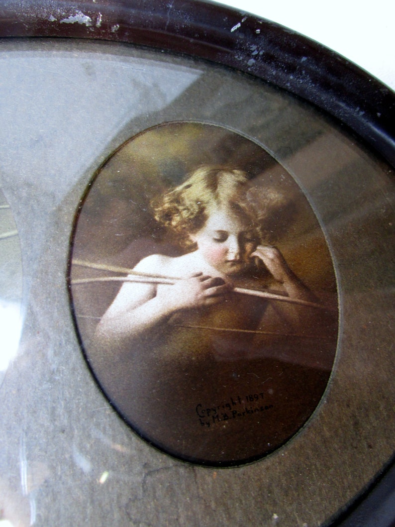 K&ouml;nnte beinhalten: Ein Vintage-Ovalrahmen mit einem Druck einer jungen Blondine, die einen Bogen und Pfeil h&auml;lt. Der Druck ist in Sepiat&ouml;nen gehalten und tr&auml;gt unten den Text "Copyright 1897 by H.B. Perkins".