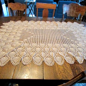 May include: Cream-colored, oval-shaped lace tablecloth with a detailed, openwork design. The edges feature a repeating heart-shaped pattern. The tablecloth is placed on a wooden table, showcasing its intricate craftsmanship.