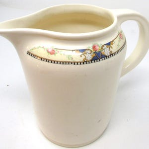 May include: Cream ceramic pitcher with a curved spout and a large handle. The pitcher has a decorative band with floral and geometric patterns in pink, blue, and gold. A classic piece of kitchenware, ideal for serving.