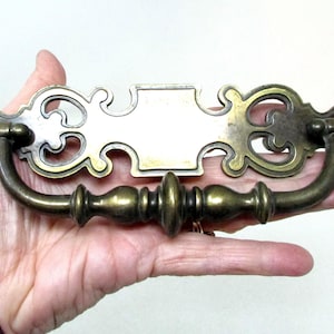 May include: Ornate brass drawer pull with a decorative design. The pull features a curved handle and decorative end pieces. The center has a rectangular plate. This vintage hardware is ideal for antique furniture restoration projects.
