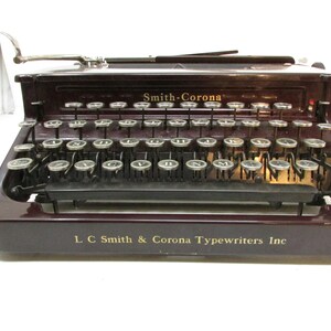 May include: A vintage Smith-Corona typewriter in a deep burgundy finish. The keys are silver and arranged in a standard QWERTY layout. The text "Smith-Corona" and "L C Smith & Corona Typewriters Inc" are visible on the machine.