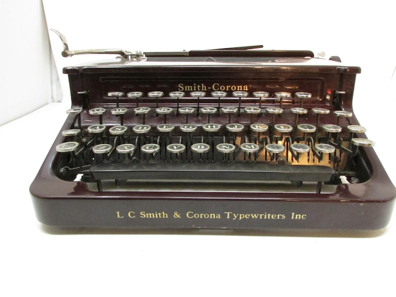 May include: A vintage Smith-Corona typewriter in a deep burgundy finish. The keys are silver and arranged in a standard QWERTY layout. The text "Smith-Corona" and "L C Smith & Corona Typewriters Inc" are visible on the machine.