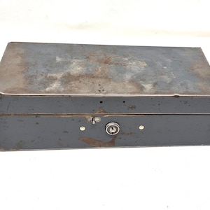 May include: A rectangular, gray metal box with a hinged lid and a lock. The box has a weathered appearance with rust-colored patches on the top. The lock is centered on the front, with two small rivets on either side.