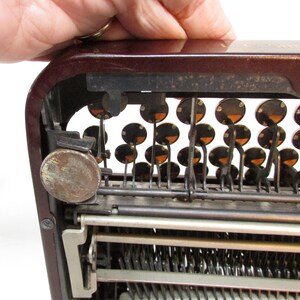 May include: Close-up of a vintage typewriter with a maroon body and black keys. The keys are round with a dark center and a lighter orange ring. The metal components show signs of age and use.