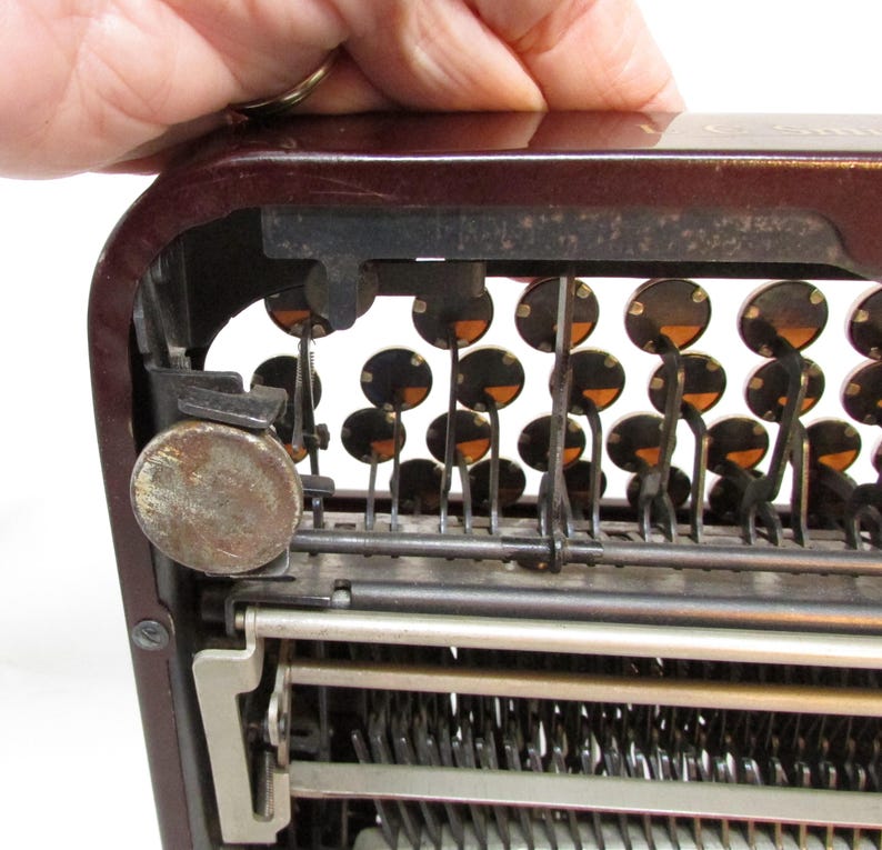 May include: Close-up of a vintage typewriter with a maroon body and black keys. The keys are round with a dark center and a lighter orange ring. The metal components show signs of age and use.
