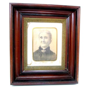 May include: A framed portrait of an elderly woman. The portrait is in a rectangular frame with a dark wood finish and a gold-coloured inner border. The woman is wearing a dark-coloured garment.