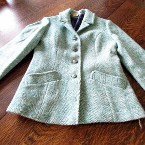 May include: A long-sleeved blazer in a light teal and white herringbone pattern. The jacket features a notched lapel, four dark buttons down the center front, and two horizontal welt pockets at the waist. The interior is lined with dark fabric.